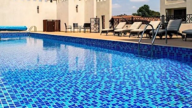 Una piscina con mosaicos azules, sillas de descanso y un edificio de fachada blanca y detalles decorativos.