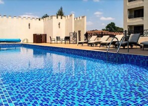 A swimming pool with blue mosaic tiles, lounge chairs, and a building with a white facade and decorative elements. - Moonvalley - View Palace Apartment (Nairobi)