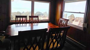 A dining area with a table and chairs, large windows with a view of the ocean and rocks, and wooden walls. - Oak Harbor Lake Erie Beach Front Cabin 3Bed 1Bath (Oak Harbor)