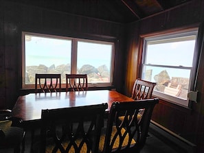 A dining area with a table and chairs, large windows with a view of the ocean and rocks, and wooden walls.
