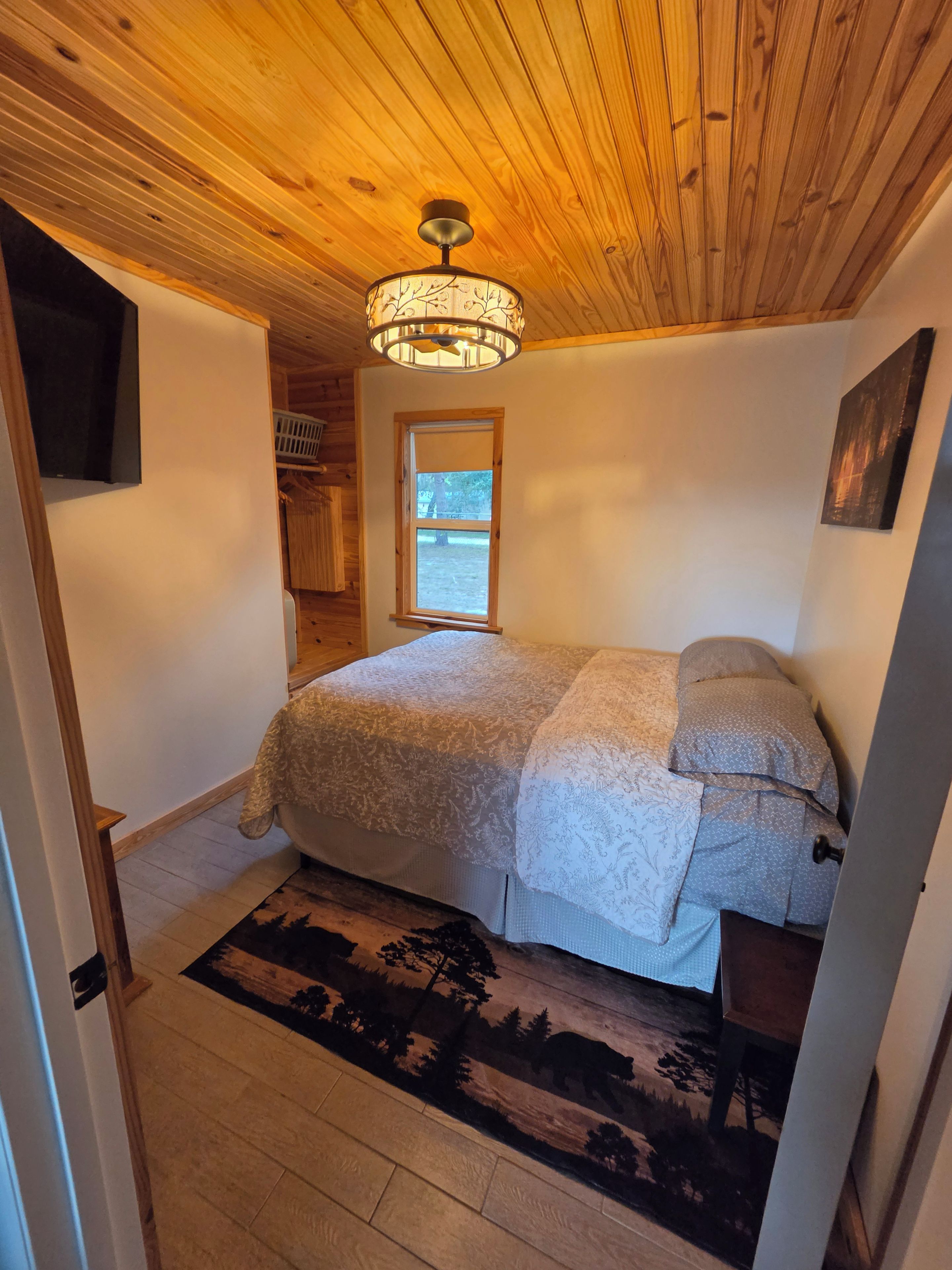 A bedroom with a wooden ceiling, a bed with patterned bedding, a window, and a door.