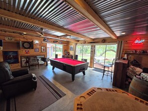 A pool table in a room with wooden walls, a neon sign, and a view of the outdoors.