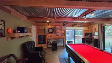 A pool table with a red felt surface, a dartboard, and a billiards cue rack.