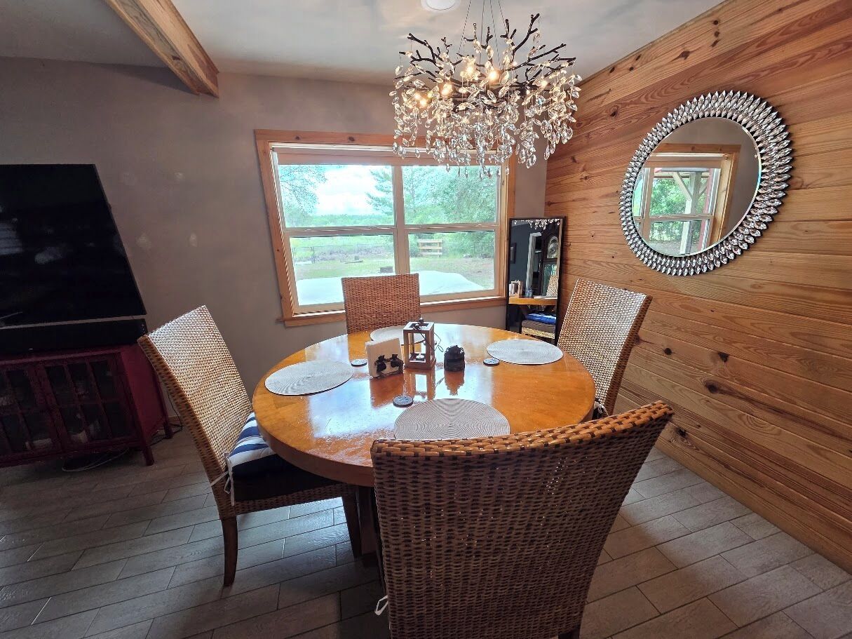 A dining area with a round table, wicker chairs, a chandelier, and a wooden wall with a mirror.