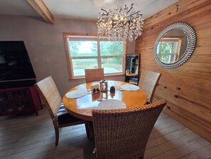 Un coin repas avec une table ronde, des chaises en osier, un lustre et un mur en bois agrémenté d’un miroir.