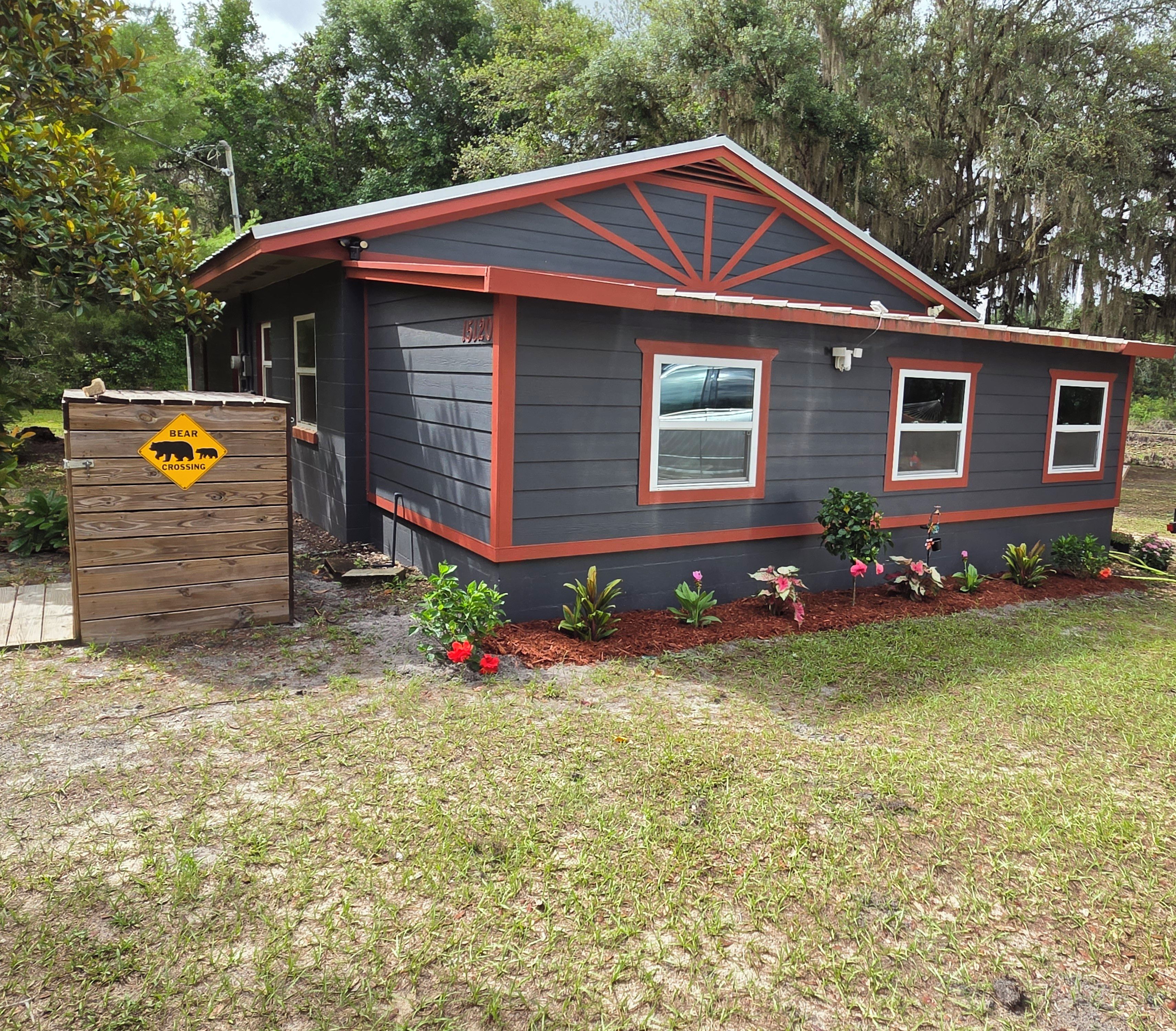 A small house with a dark exterior and red trim, a bear crossing sign, and a well-maintained garden with various plants and flowers.