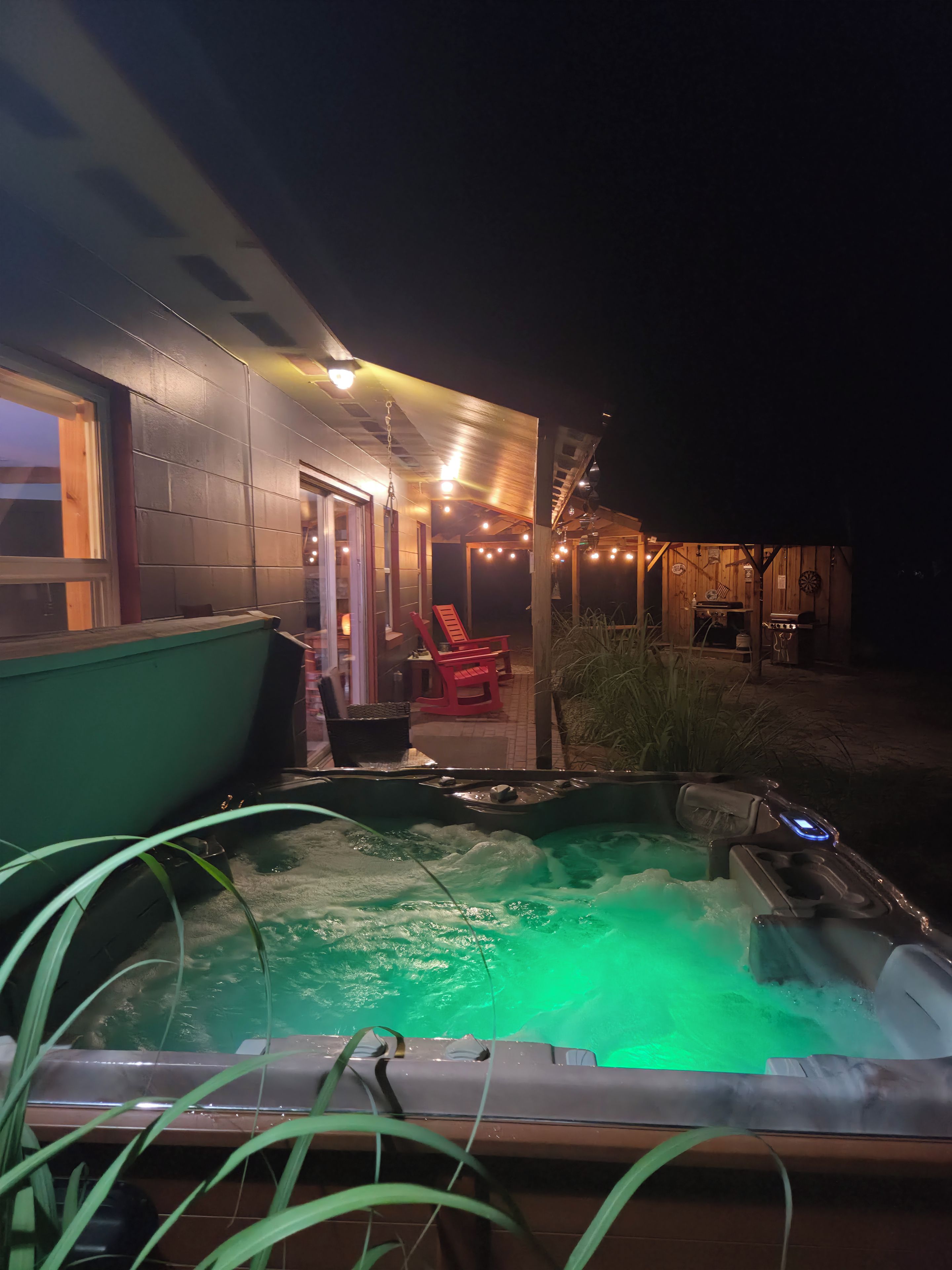Un jacuzzi dont l’intérieur est éclairé en vert, un patio couvert avec des chaises rouges, et un bâtiment dont on peut voir la porte et les fenêtres.