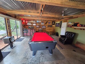 A pool table in a room with wooden beams, a bar area, and a view through large windows.