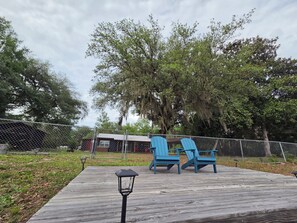A wooden deck with two blue Adirondack chairs, a lantern, and a fenced area with trees and a building in the background.