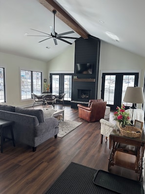 A spacious living room with a fireplace, a ceiling fan, and large windows.