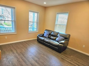 A room with a leather sofa, wooden flooring, and large windows with blinds.