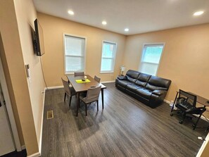 A living room with a dining table, a black leather sofa, and a TV mounted on the wall.