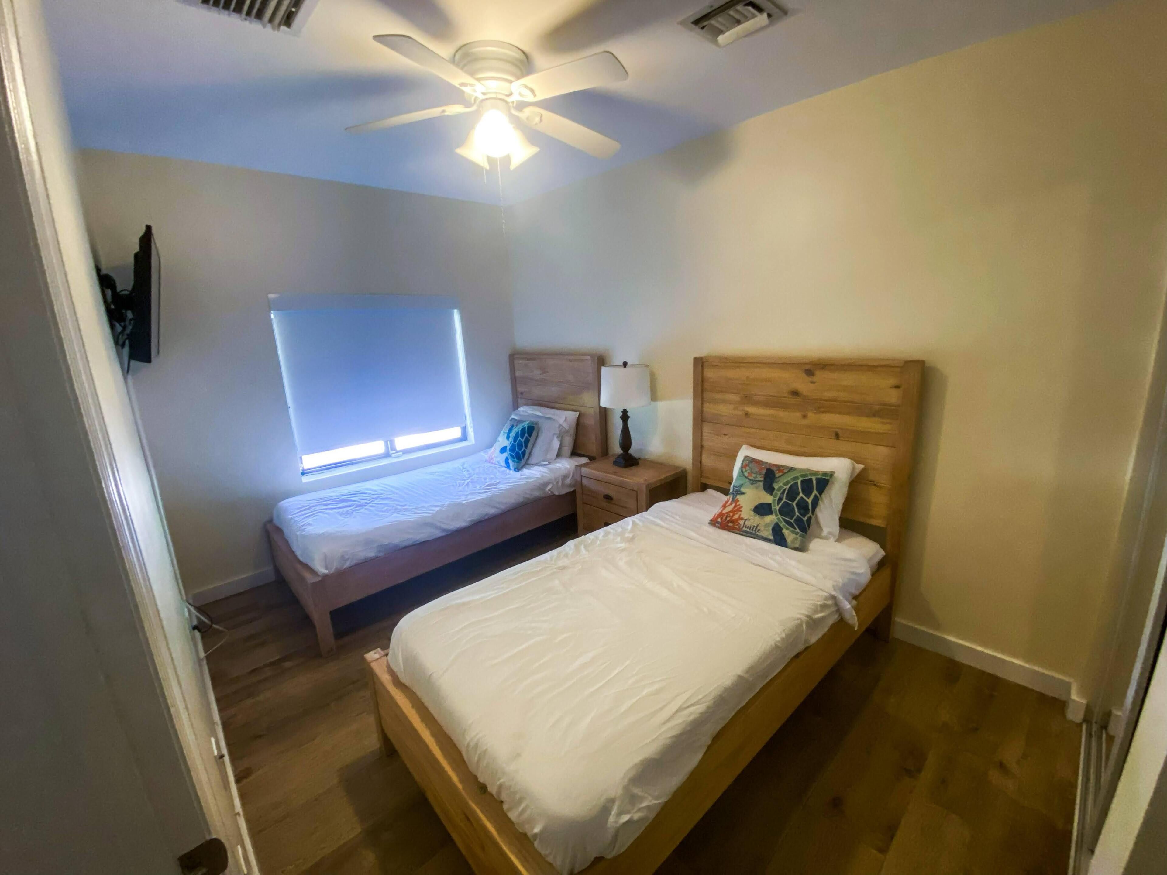Une chambre avec deux lits, un ventilateur de plafond, une fenêtre avec des stores et une table de chevet avec une lampe.