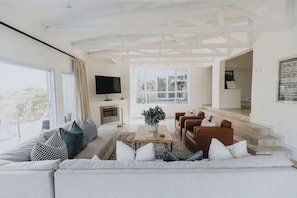 A modern living room with a sofa, coffee table, and fireplace. There is a TV mounted on the wall and a staircase leading to another level. - Stunningly decorated home with breathtaking views! (Helensvale)
