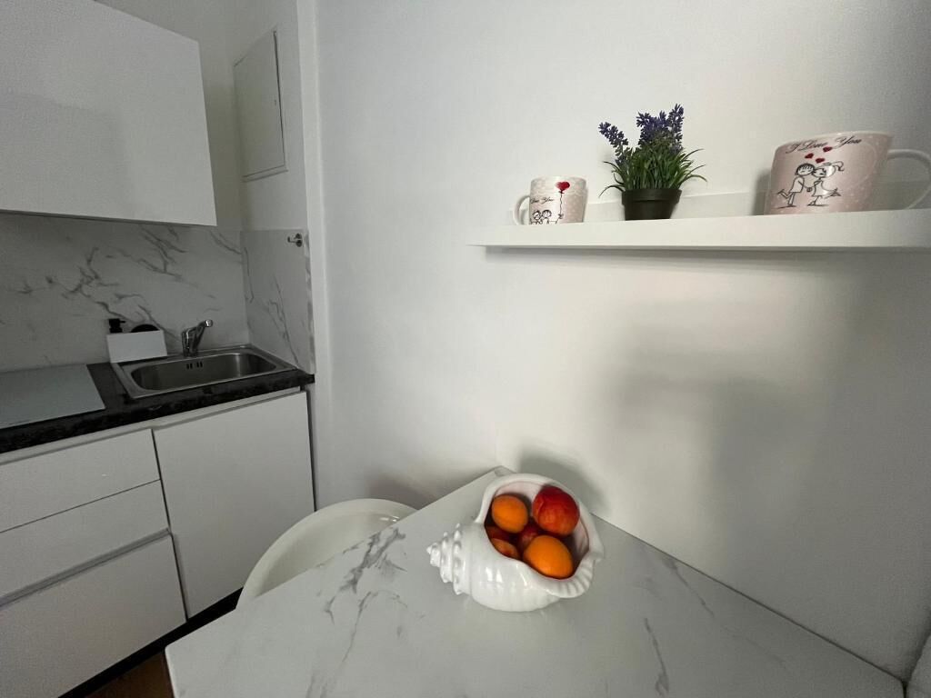 A kitchen with white cabinets, a marble countertop, a sink, a white table with a fruit bowl, and a shelf with decorative items.