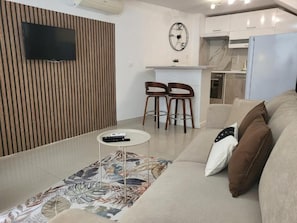 Olohuoneessa on sohva, sohvapöytä, puiseen seinään asennettu televisio sekä keittiöalue, jossa on jääkaappi ja kello.