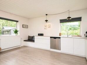 A modern kitchen with white cabinets, a built-in dishwasher, a bench with cushions, pendant lighting, and a view of greenery outside. - 5 Star Holiday Home in Farvang (Farvang)