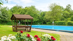 Eine hölzerne Laube mit einer Bank, einem Schwimmbecken und Blumenbeeten mit roten und weißen Blüten.