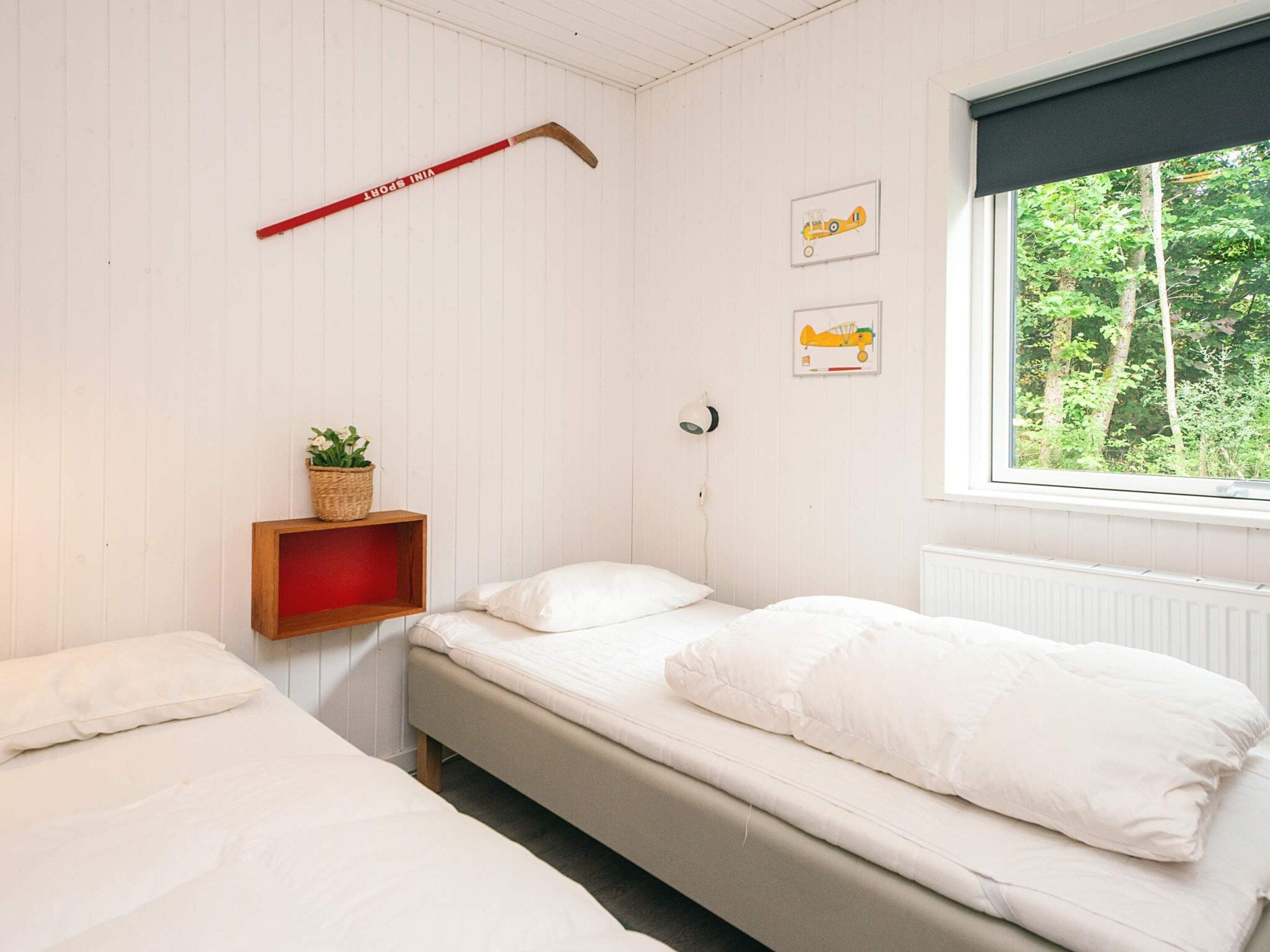 Una habitación con dos camas, una ventana que da a árboles, una lámpara de pared y un palo de hockey rojo.