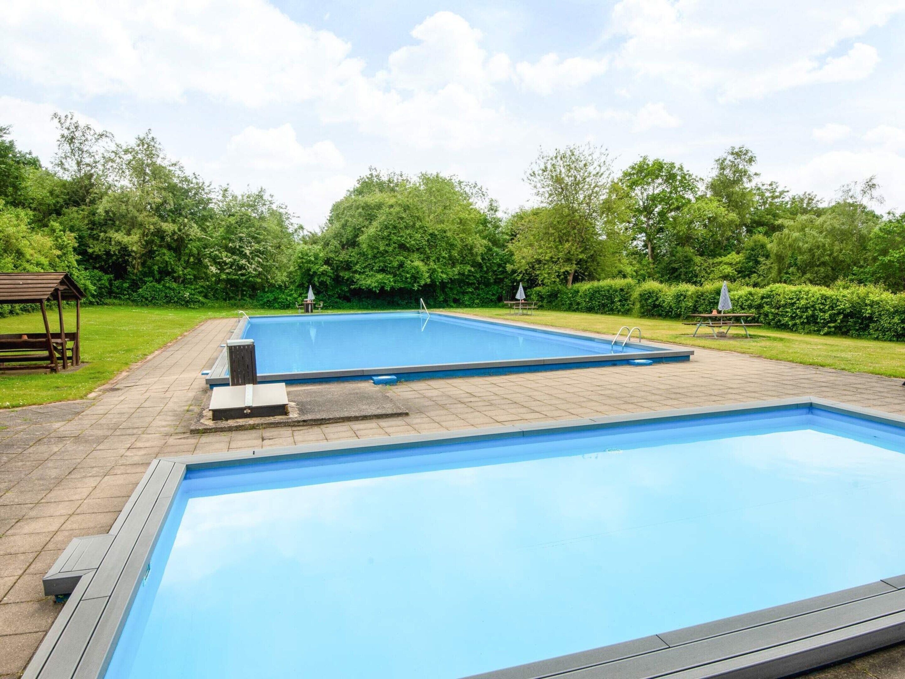 Une piscine extérieure rectangulaire, dotée d’une terrasse carrelée et entourée de verdure et d’arbres.