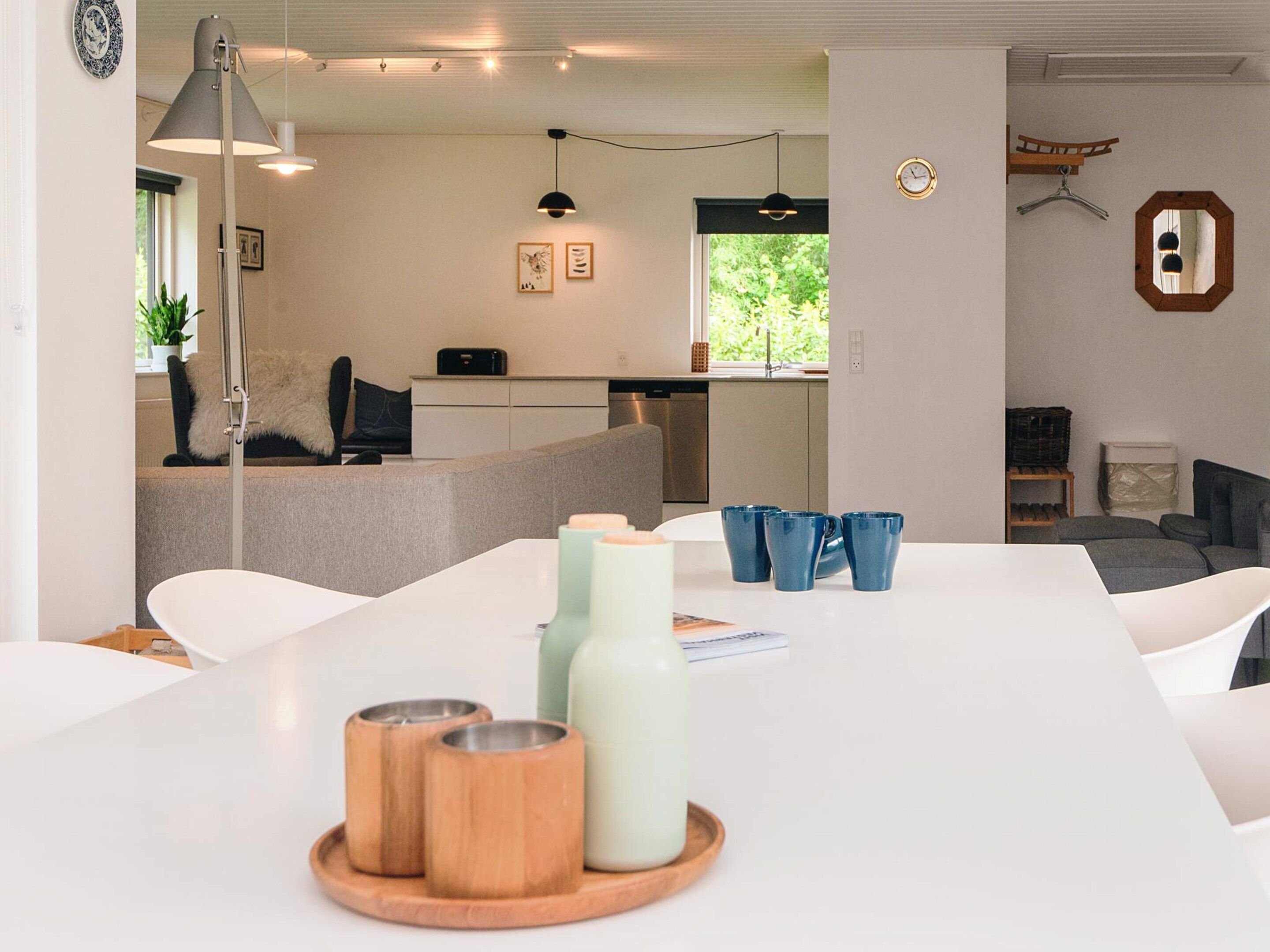 A modern living room with a white table, a sofa, and a dining area. There are decorative items on the table, and a kitchen area is visible in the background.