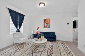Ein Wohnzimmer mit einem weißen Sofa, einem blauen Couche, einem runden Couchtisch mit goldfarbener Basis, einem gemusterten Teppich und einem Bild an der Wand.