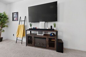 A living room with a flat-screen TV mounted on the wall, a wooden entertainment unit below, a yellow towel on a stand, and a potted plant. - Blue Yard Homes • 4BR Near JBSA Randolph • Hot Tub & PS5 (Converse)