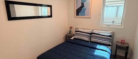 Un dormitorio con cama, mesita de noche, ventana y un espejo instalado en la pared.