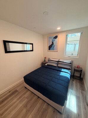 Ein Schlafzimmer mit einem Bett, einem Nachttisch, einem Fenster und einem an der Wand befestigten Spiegel.