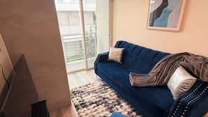 A living room with a blue sofa, a small ottoman, a fluffy rug, a TV stand, and a sliding glass door leading to a balcony. - Chic Urban Luxury Retreat 1Bedroom Apartment (Los Angeles)
