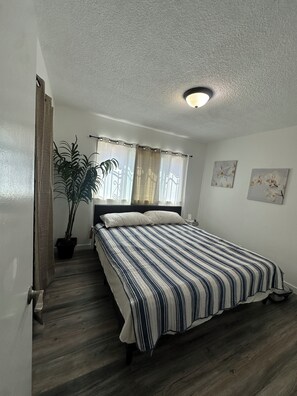 Ein Schlafzimmer mit einem Bett, einer Topfpflanze, einem Fenster mit Vorhängen und zwei an der Wand befestigten Bildern.