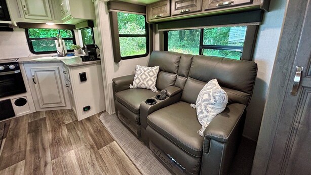 A compact RV interior with a kitchenette, a sofa with cushions, and windows providing views of greenery.