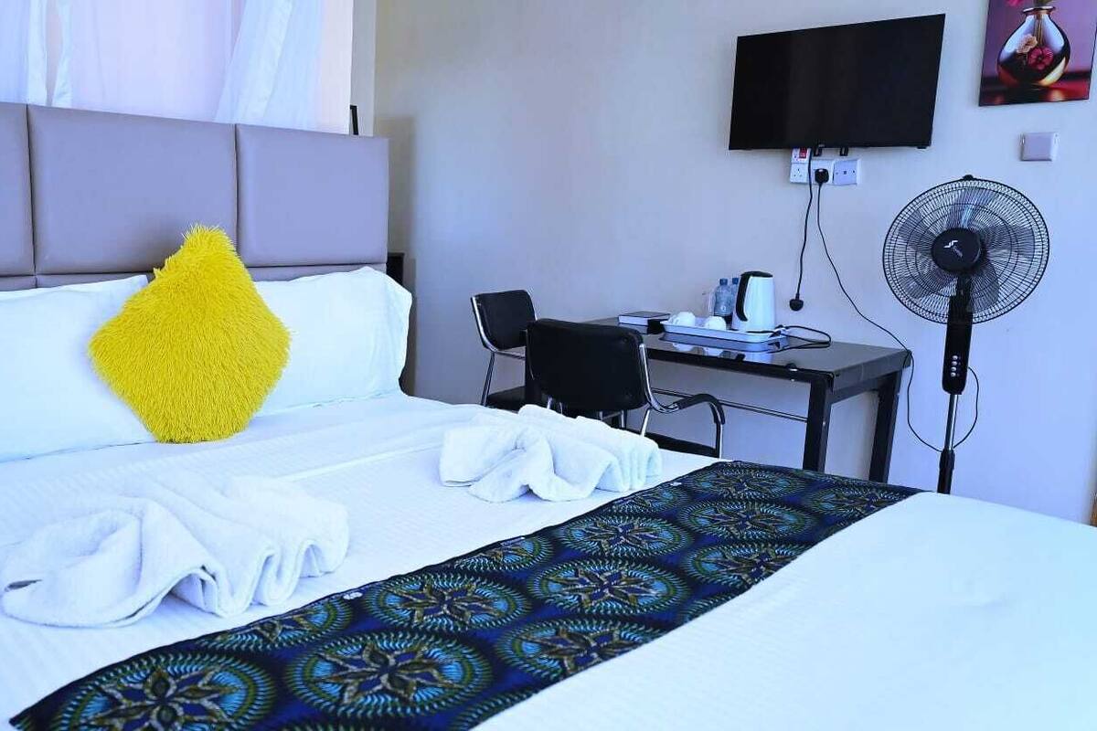 A hotel room with a bed, a desk, a chair, a TV, and a standing fan.