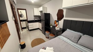 Quarto moderno com cama, mesa de cabeceira e pequena cozinha equipada com frigorífico e armários.