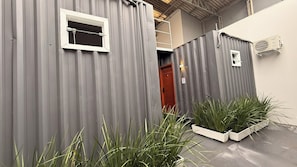 A corrugated metal structure with a window and a door, surrounded by potted plants. - LOFT127 - 04 (Itajaí)