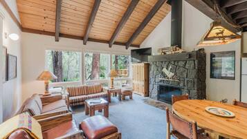 A cozy living room with a stone fireplace, wooden ceiling beams, and a large window overlooking a forest.