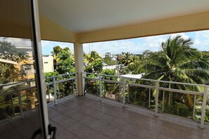 A balcony with a tiled floor, white railing, and a view of palm trees and buildings. - Tropical villa with a large pool, lush gardens, spacious rooms, and privacy. (Pointe Aux Cannioner)