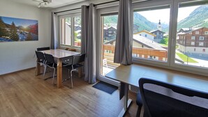 Ein Zimmer mit Holzboden, einem Esstisch mit Stühlen, einem Deckenventilator und großen Fenstern mit Vorhängen.