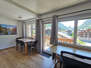 Ein Zimmer mit Holzboden, einem Esstisch mit Stühlen, einem Deckenventilator und großen Fenstern mit Vorhängen.