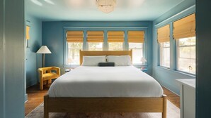 Une chambre à coucher avec un lit en bois, deux lampes de chevet, une chaise et de grandes fenêtres équipées de stores.