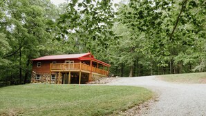 A cabin with a stone foundation, wooden deck, and a red roof, surrounded by a forested area. - Lovely 4-bedroom cabin with AC in pleasant Shoals (Shoals)