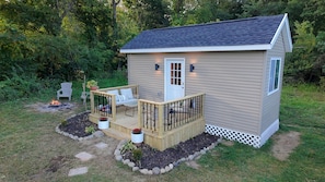 A small, single-story wooden cabin with a deck, a fire pit, and a garden area.