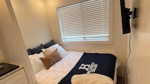 A bedroom with a bed, a window with blinds, a bedside table, and a wall-mounted TV. - Tiny House near Hillsdale College (Hillsdale)