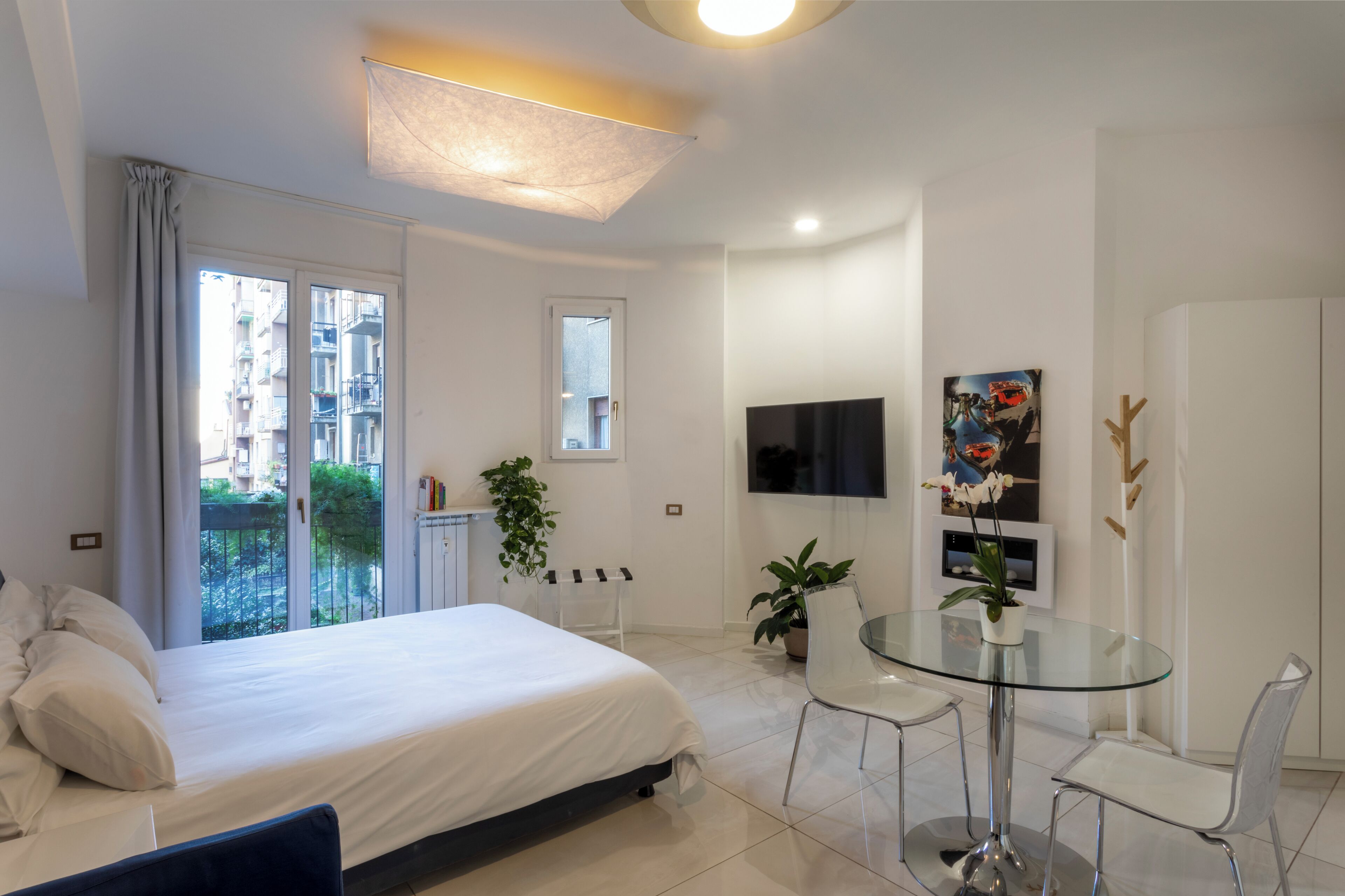 A modern hotel room with a bed, a glass dining table, chairs, a TV, and a balcony with a view of buildings.