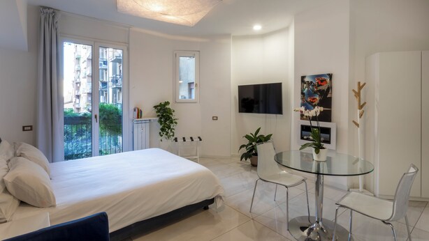 Ein modernes Hotelzimmer mit Bett, einem Glastisch, Stühlen, einem Fernseher und einem Balkon mit Blick auf Gebäude.