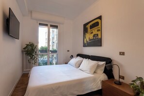 Ein Schlafzimmer mit Bett, Nachttisch, Lampe, Fenster, Heizkörper, Pflanze und einem an der Wand befestigten Fernseher.