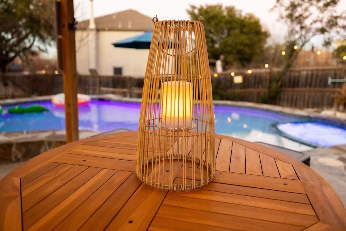 Une table en bois avec une bougie dans un photophore tressé, une piscine en arrière-plan et une clôture illuminée.