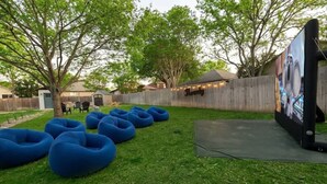 A backyard with a wooden fence, a tree, and blue bean bags arranged on the grass. - Heated Pool • Game Rm • Theater • Firepit • Pets (San Antonio)
