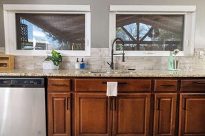 A kitchen with two windows, a dishwasher, wooden cabinets, and a countertop with various items. - Heated Pool • Game Rm • Theater • Firepit • Pets (San Antonio)