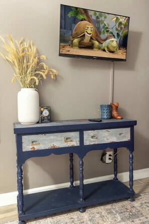 A wall-mounted television displaying a cartoon character, a decorative vase with dried plants, and a vintage-style blue console table with drawers. - Heated Pool • Game Rm • Theater • Firepit • Pets (San Antonio)